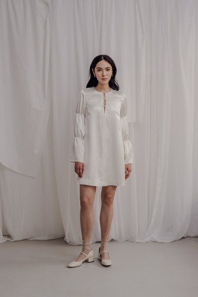 Die Braut trägt ein kurzes Kleid, welches gerüschte Ärmel hat und am Ausschnitt mit drei kleinen Schleifen besetzt ist