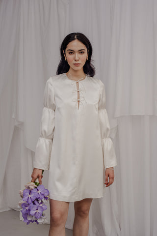 Die Braut trägt ein kurzes Kleid, welches gerüschte Ärmel hat und am Ausschnitt mit drei kleinen Schleifen besetzt ist